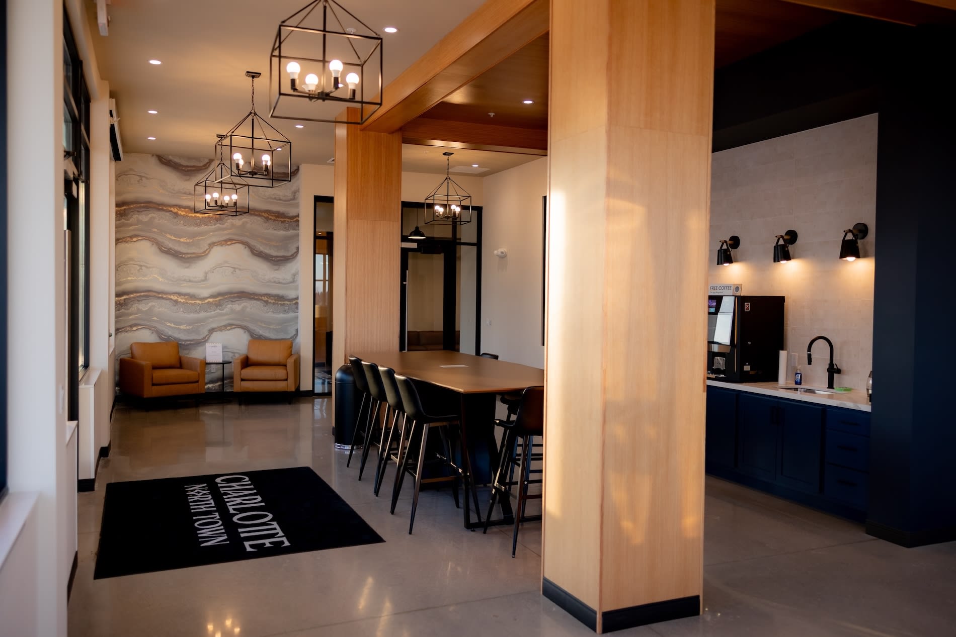 Open-concept common area with wood dining table, black chairs, orange lounge seating, geometric pendant lights, and navy kitchen with black countertops.