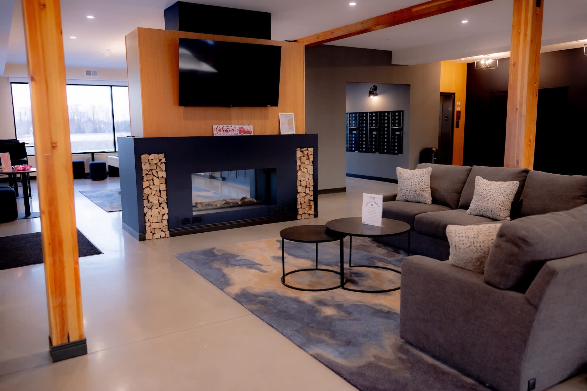 Open-concept lounge with gray sectional sofa, linear fireplace with wood storage, wall-mounted TV, round coffee tables, and large windows overlooking the city.