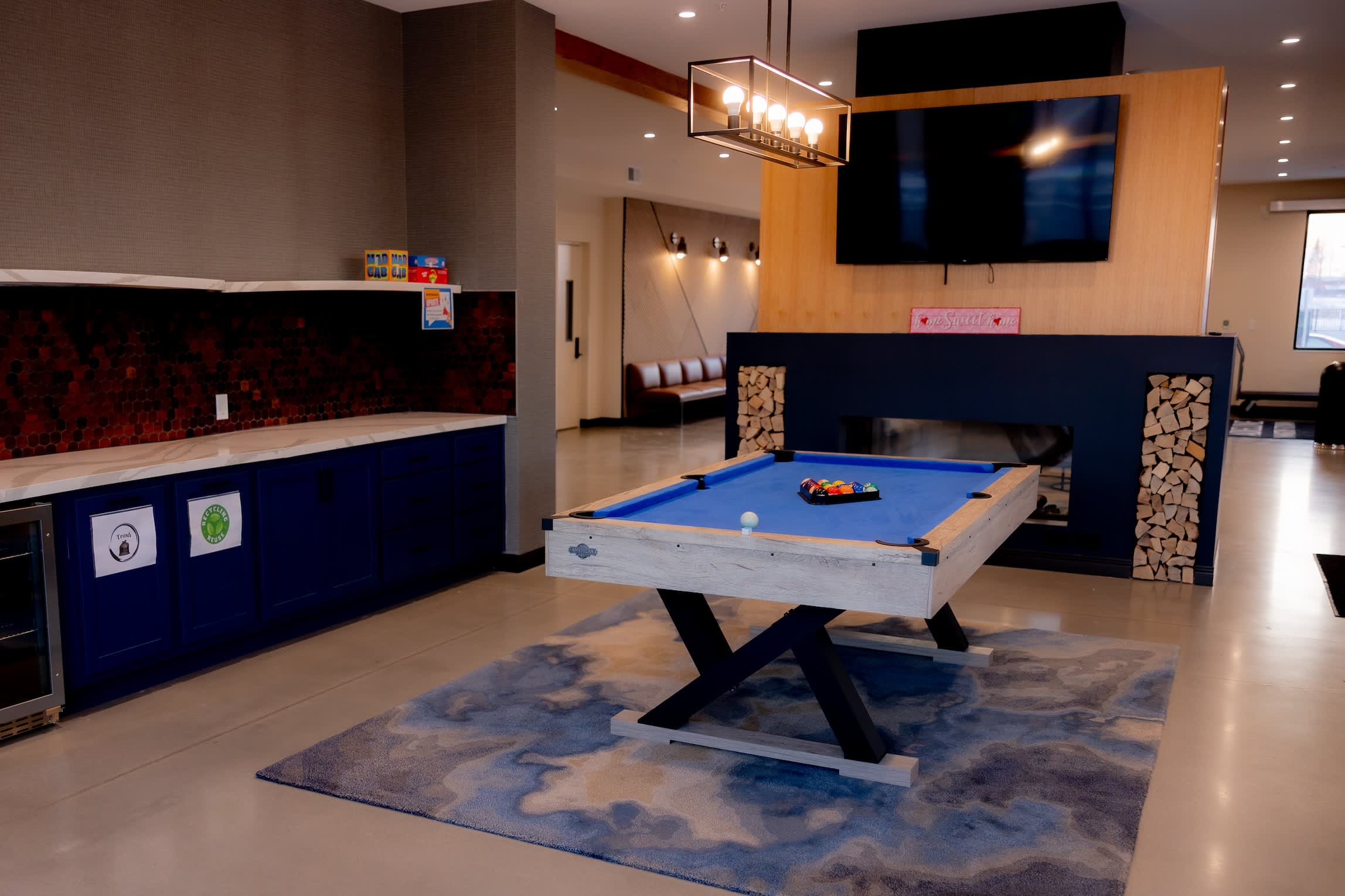 Recreation room with blue pool table, navy kitchen cabinetry with red backsplash, wall-mounted TV, and fireplace with wood storage.