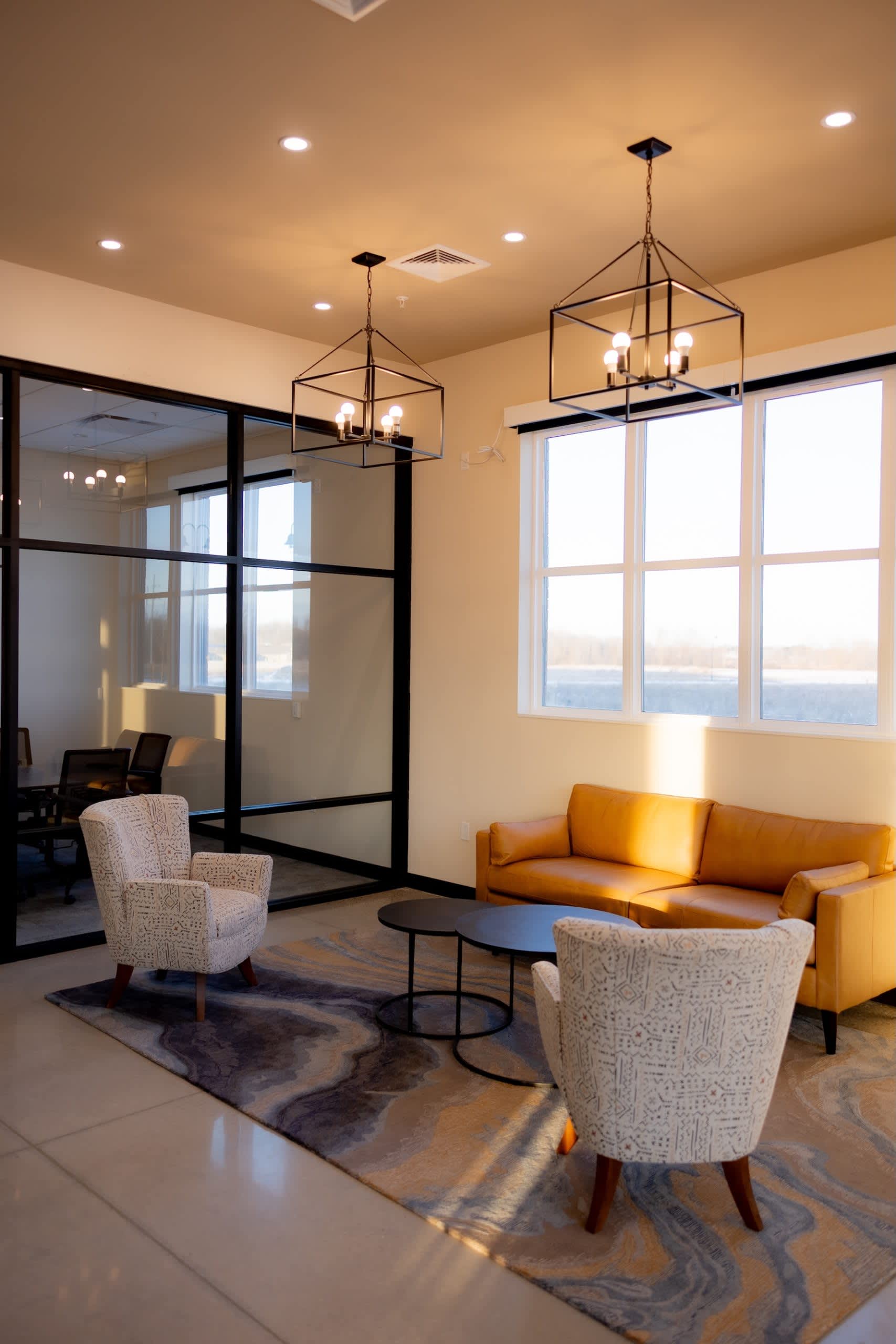 Modern apartment lounge with orange sofa, cream armchairs, black-framed glass partition, and large windows overlooking water.