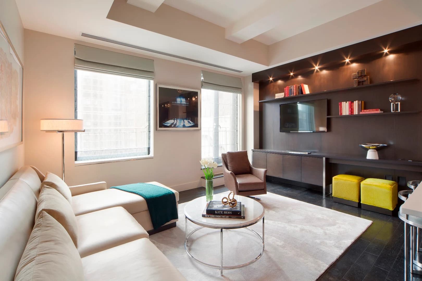 Modern living room with cream sofa, dark brown accent wall with built-in shelving and TV, round coffee table, and large windows with city views.