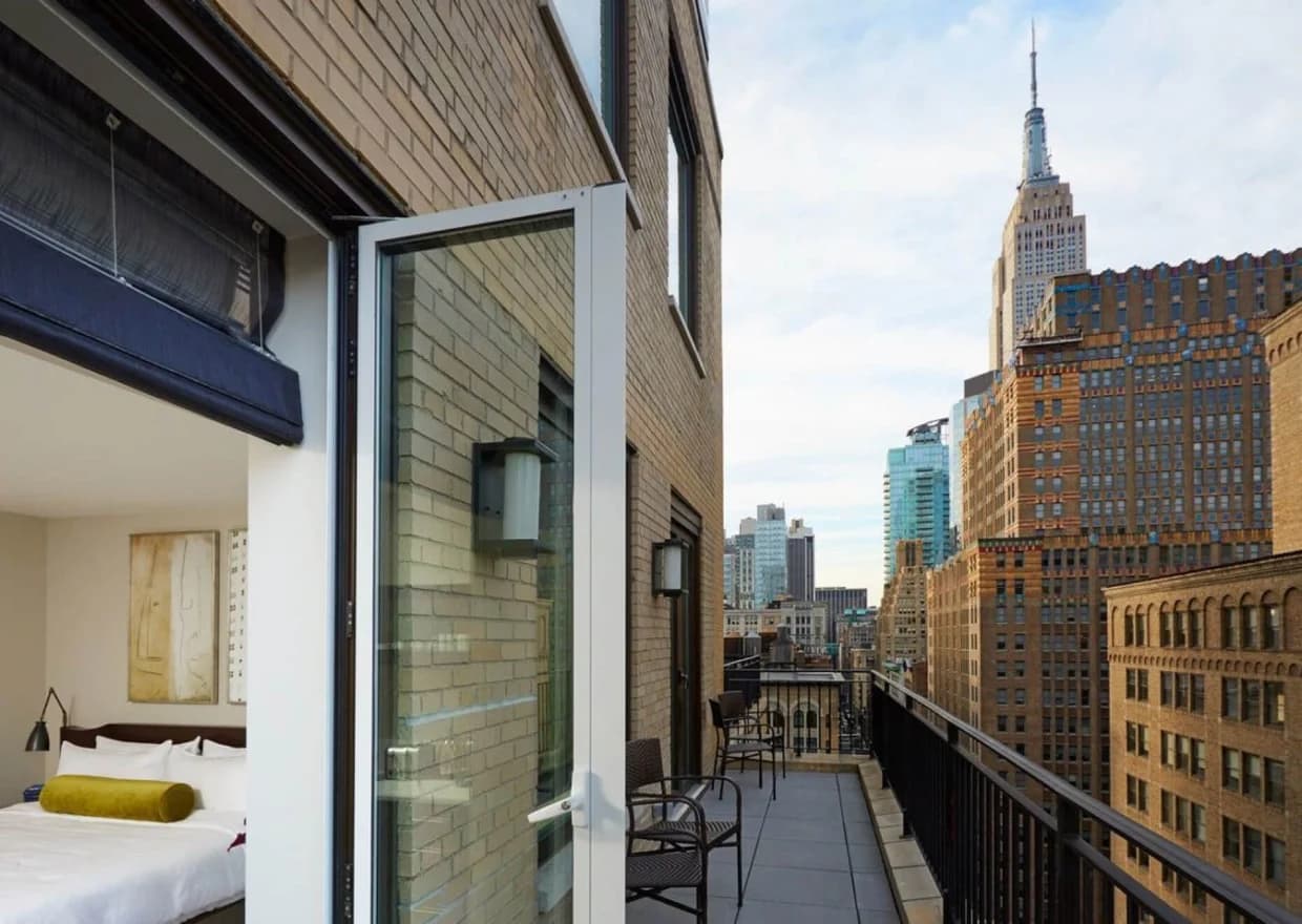 Bedroom with white bedding and yellow accent pillow opening onto a narrow terrace with seating, facing Empire State Building and surrounding skyscrapers.