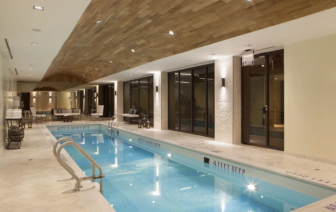 Indoor swimming pool with blue water, metal handrails, wood-beam ceiling, recessed lighting, and dark glass doors along cream-colored walls.