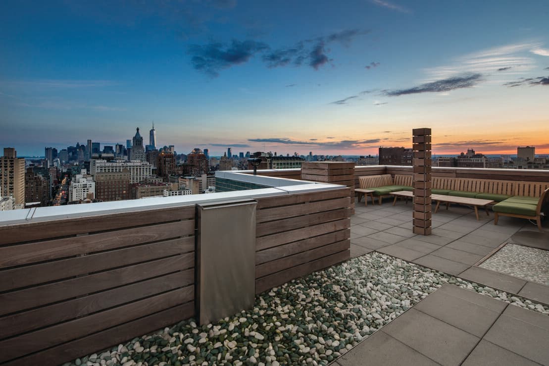 Rooftop terrace with wooden privacy screens, lounge seating, gravel planters, and expansive Manhattan skyline at sunset.