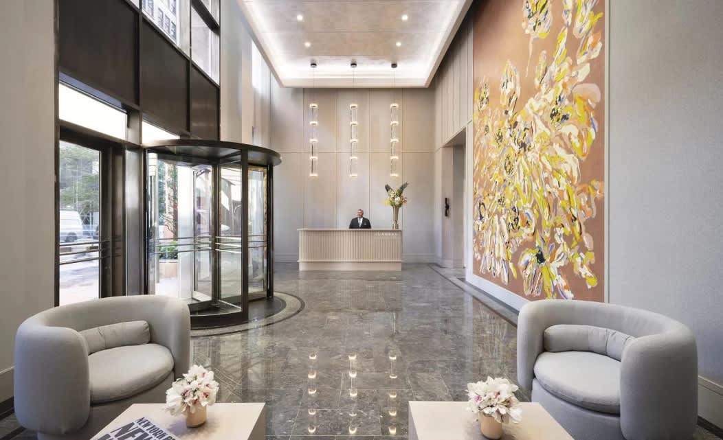 Modern lobby with gray curved armchairs, polished marble floor, reception desk, glass entrance doors, and large yellow abstract artwork on wall.