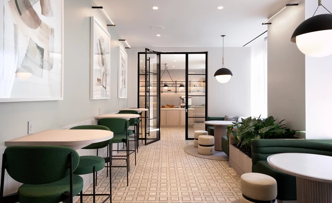 Modern common area with green upholstered chairs at round tables, geometric floor pattern, black-framed glass doors, pendant lighting, and potted plants.