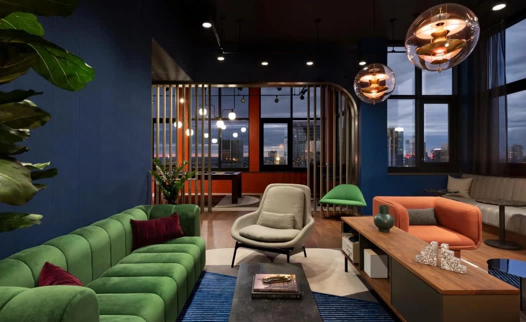 Modern lounge with green curved sofa, orange armchair, wooden table, and panoramic city skyline views through large windows.