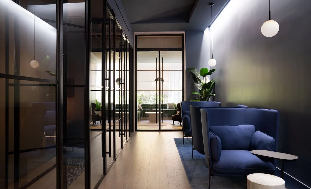 Modern hallway with black-framed glass partitions, navy blue armchair, pendant lighting, and glass doors opening to a terrace with city views.
