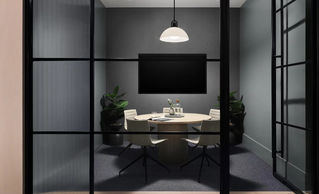 Modern conference room with round wood table, four black chairs, pendant light, potted plants, and black-framed glass walls.