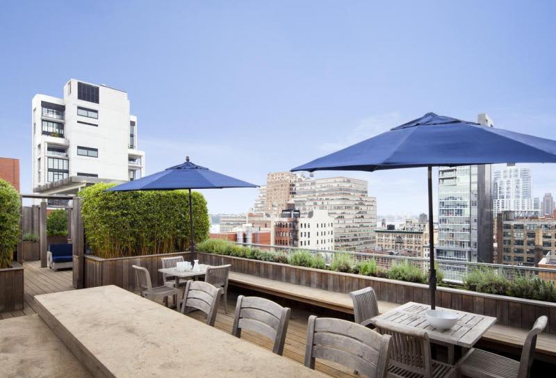 Rooftop terrace with wooden dining tables, gray chairs, blue umbrellas, bamboo planters, and Manhattan skyline views.