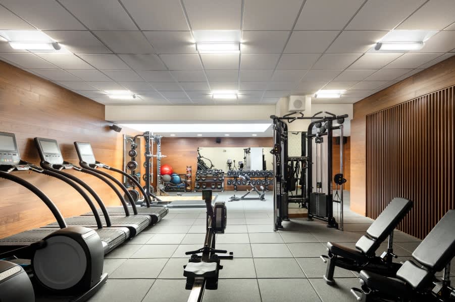 Fitness center with treadmills on left, stationary bikes in center, weight machines and strength training equipment, wood-paneled walls, and bright ceiling lights.