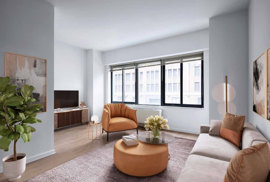Modern living room with tan leather armchair and ottoman, beige sofa, large black-framed windows overlooking city buildings, potted plant, and wood media console.