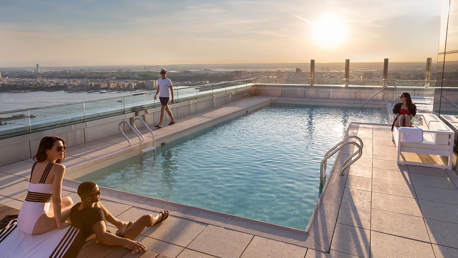 Rooftop swimming pool with glass railings, sunbathers, and expansive cityscape view at golden hour.
