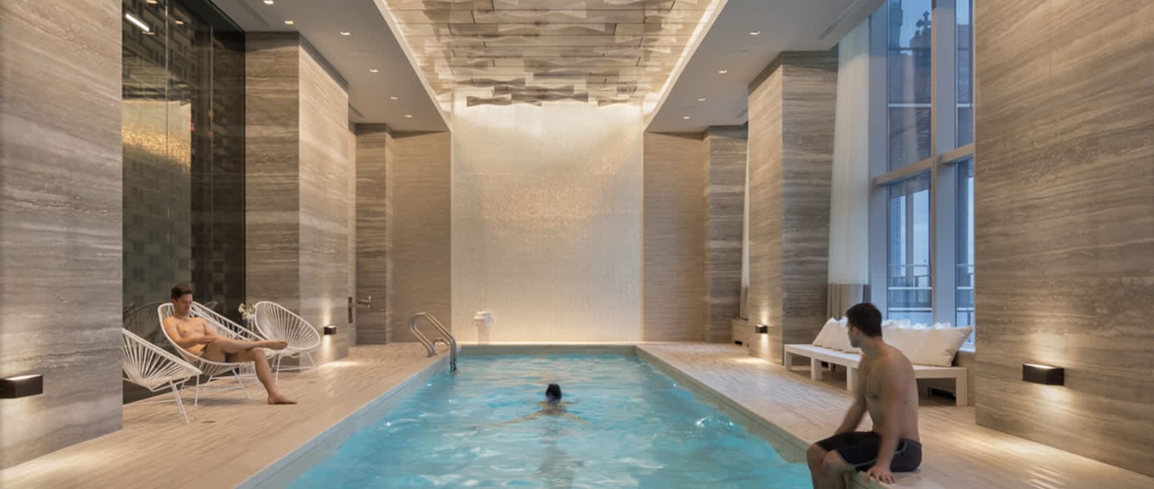 Indoor swimming pool with blue water, stone tile walls, white lounge chairs, and large windows in a modern building.