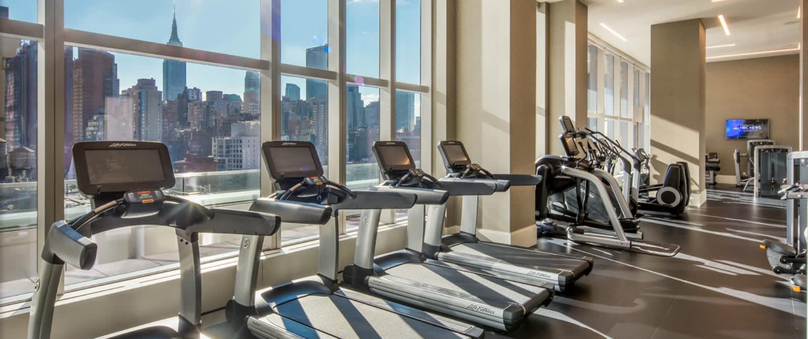 Modern gym with treadmills, stationary bikes, and elliptical machines facing floor-to-ceiling windows with views of Manhattan skyscrapers.