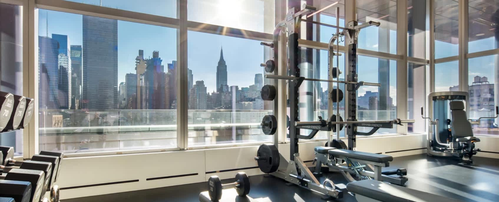 Modern gym with weight rack, dumbbells, and exercise equipment facing expansive windows with views of Manhattan skyscrapers including the Chrysler Building.
