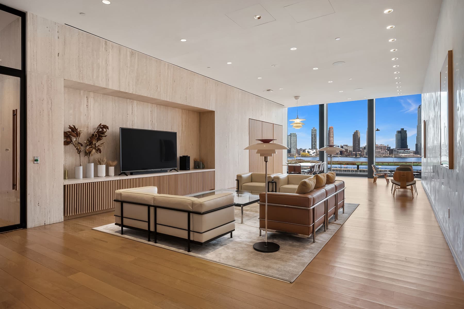 Modern living room with light wood accent wall, built-in TV, beige seating, and expansive windows framing city and water views.