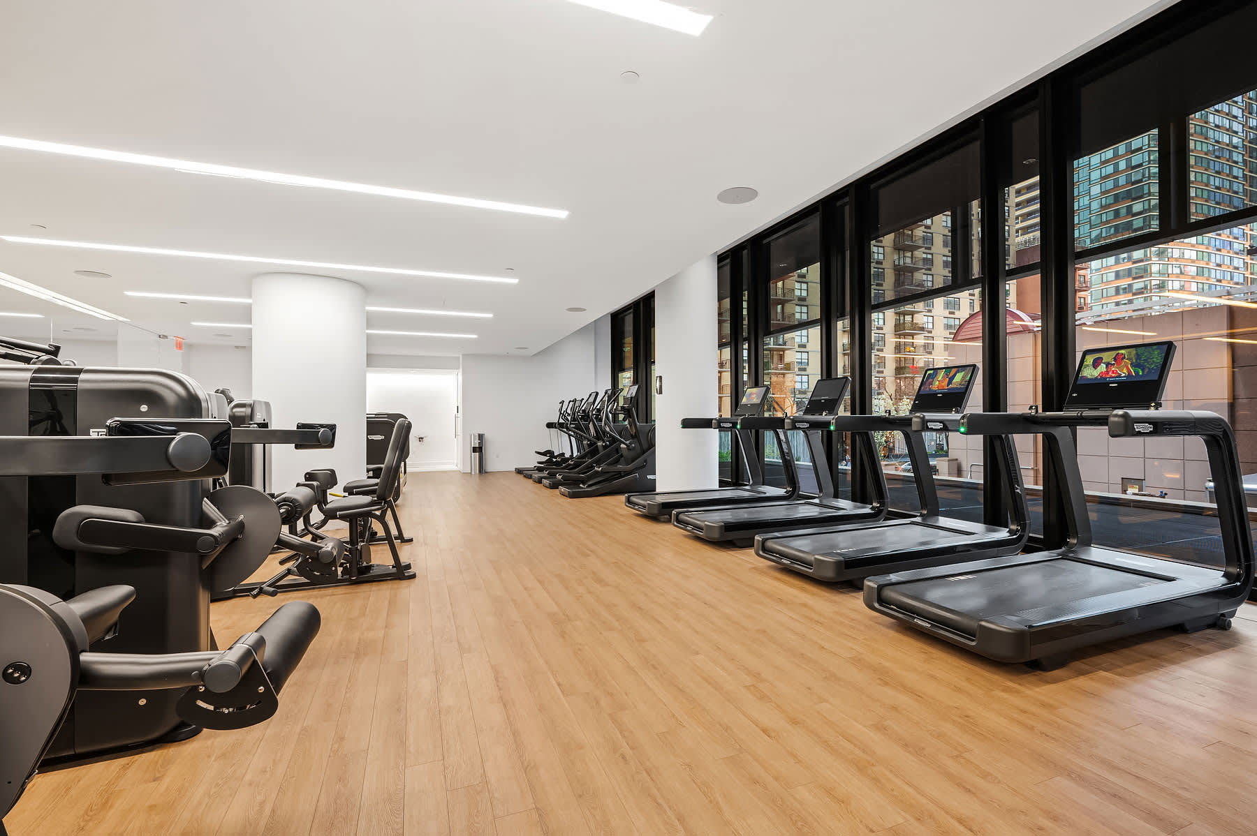 Fitness center with three treadmills, stationary bikes, and exercise equipment on wood flooring, floor-to-ceiling windows with city views.