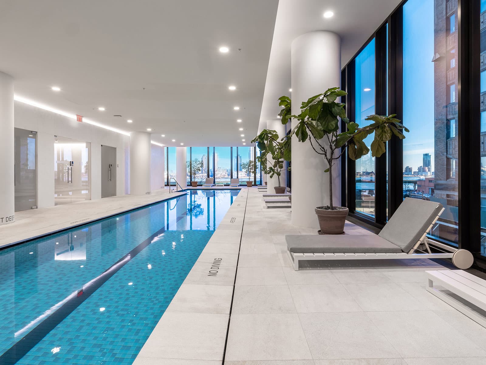 Indoor lap pool with blue tile, white deck, potted plants, lounge chair, and floor-to-ceiling windows with city views.