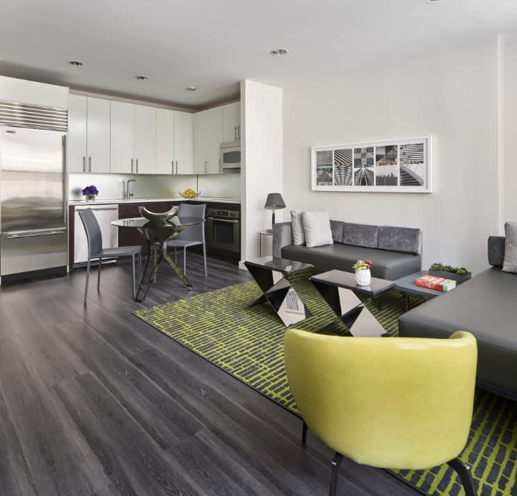 Open-concept studio with white kitchen cabinets, stainless steel refrigerator, gray sofa, yellow accent chair, and dark wood flooring.