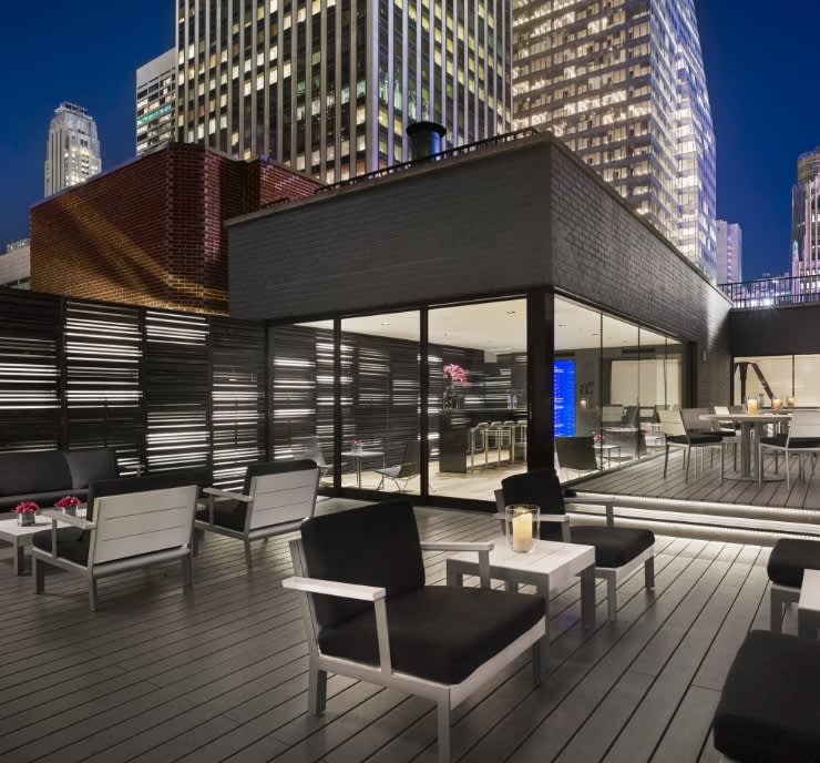 Rooftop terrace at dusk with modern lounge chairs, wooden decking, glass-walled interior space, and Manhattan skyscrapers illuminated against blue sky.