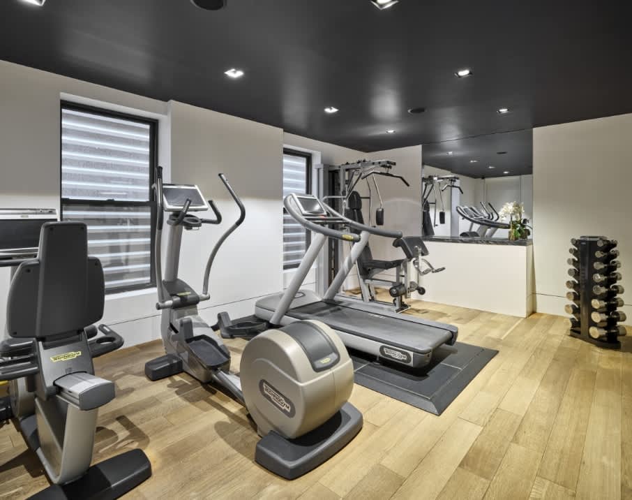 Modern fitness center with elliptical machines, treadmill, stationary bikes, dumbbell rack, and wood flooring under dark ceiling.
