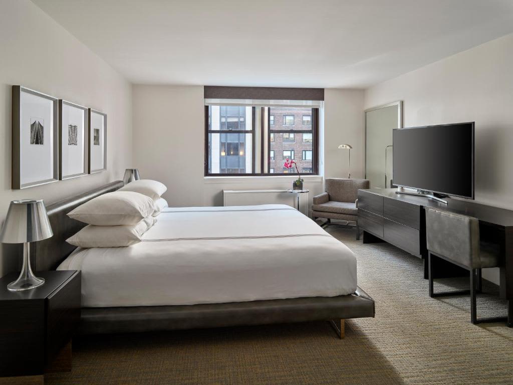 Modern bedroom with king bed, white bedding, dark wood nightstands, floor-to-ceiling windows overlooking city buildings, flat-screen TV, and seating area.