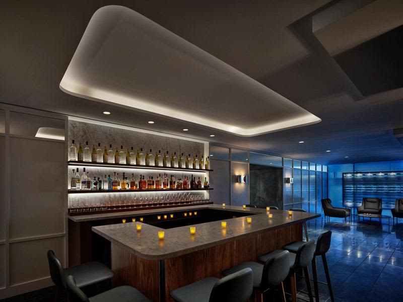 Modern bar lounge with backlit liquor shelves, dark wood counter with candles, black bar stools, and city views through floor-to-ceiling windows.