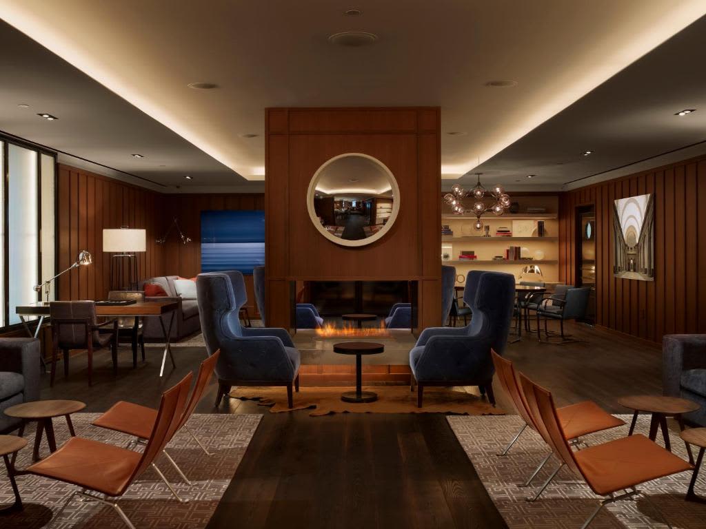 Modern lounge with wood-paneled walls, fireplace with round mirror, blue and orange mid-century chairs, and ambient ceiling lighting.