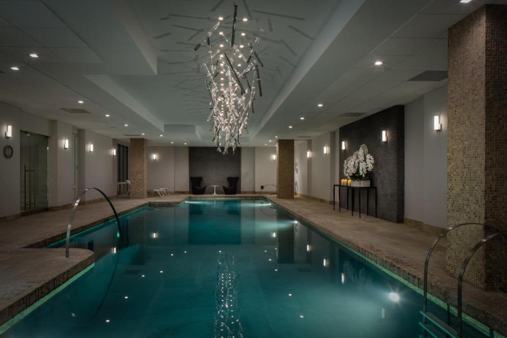 Indoor swimming pool with teal water, crystal chandelier overhead, brick columns, and recessed ceiling lights.