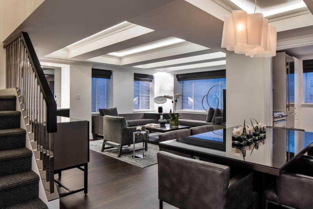 Modern living room with gray sectional sofa, black leather chairs, coffered ceiling with skylights, and large windows overlooking the city.