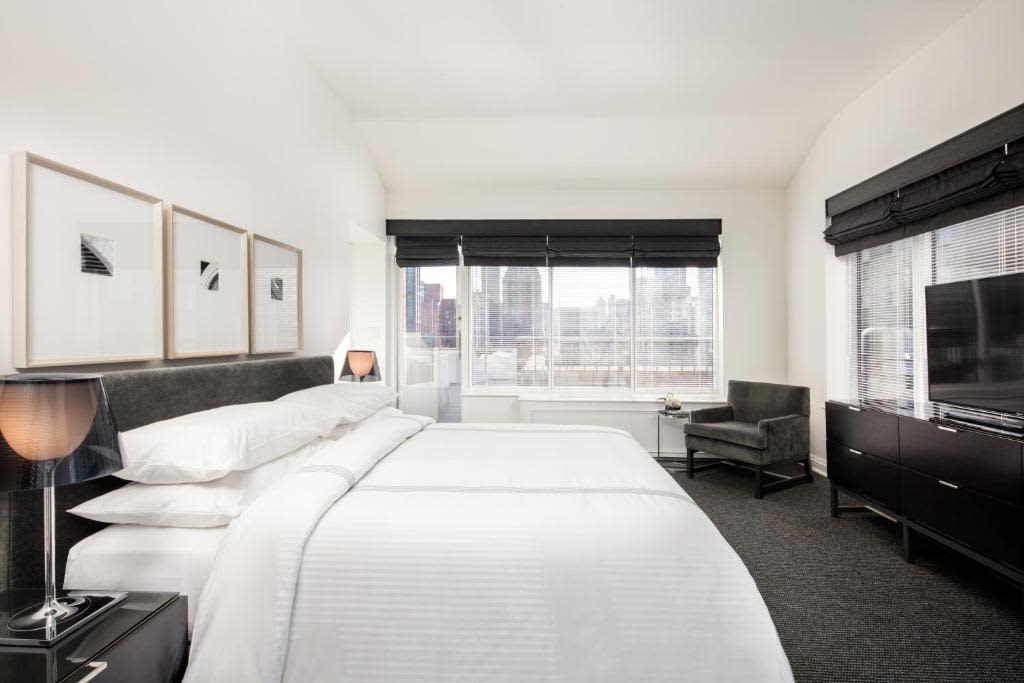 Bedroom with queen bed, dark headboard, white bedding, floor-to-ceiling windows with city views, dark accent chair, and TV on black dresser.