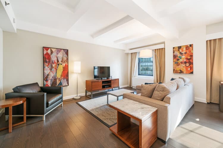 Open living room with white coffered ceiling, black leather chair, beige sofa, wooden coffee table, TV on media console, and large windows with tan curtains.