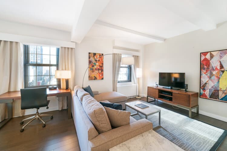 Modern living room with beige sofa, wooden media console, desk by window, and colorful abstract wall art in New York apartment.