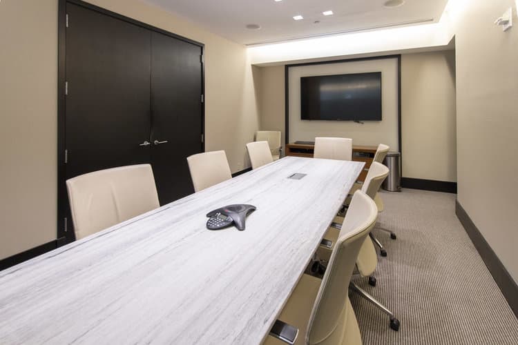 Conference room with long white table, cream upholstered chairs, wall-mounted TV, and black doors.
