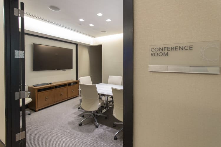 Conference room with wall-mounted TV, wooden credenza, white chairs around white table, and recessed ceiling lights.