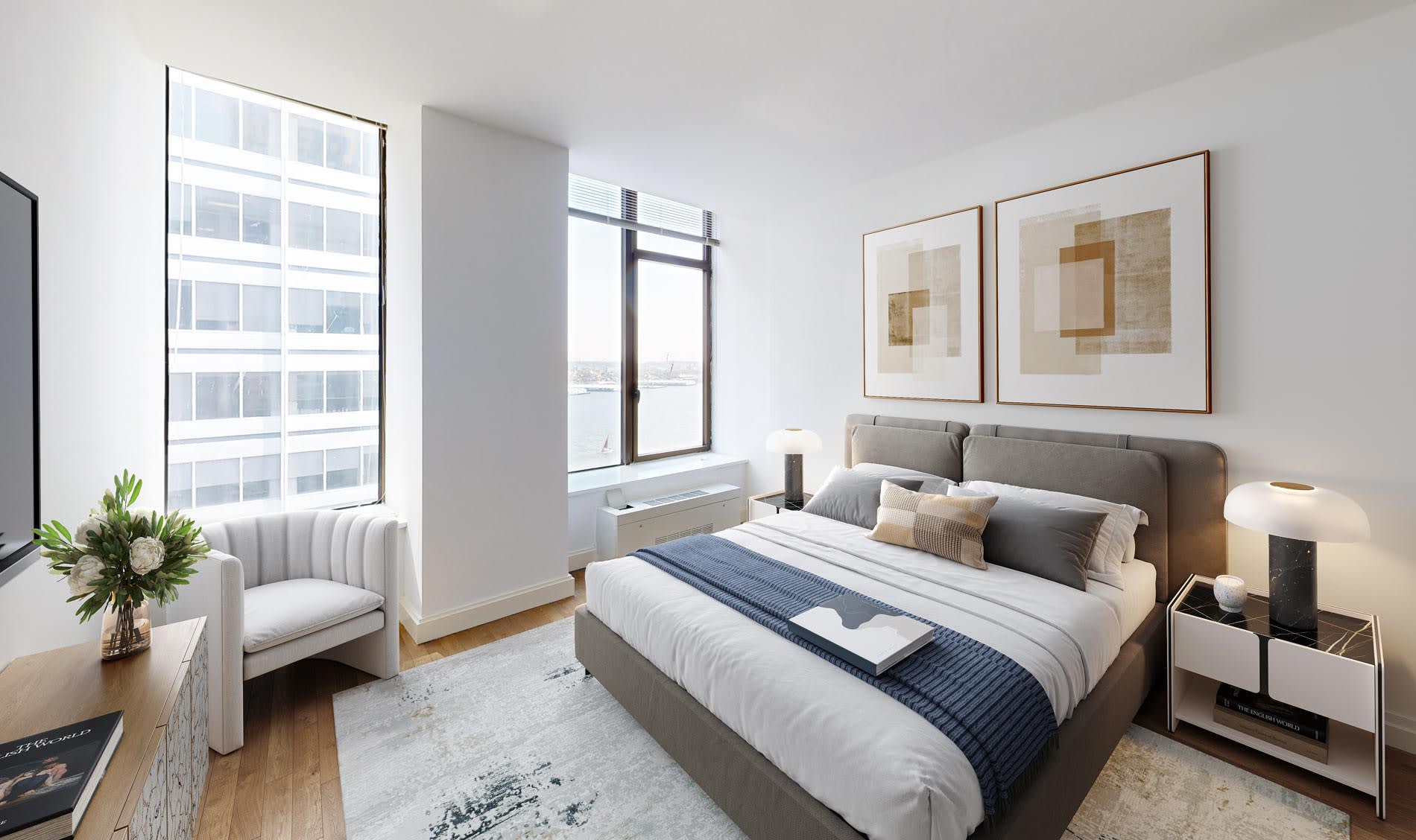 Bedroom with gray upholstered bed, two large windows with city and water views, white accent chair, framed abstract art, and hardwood floors.