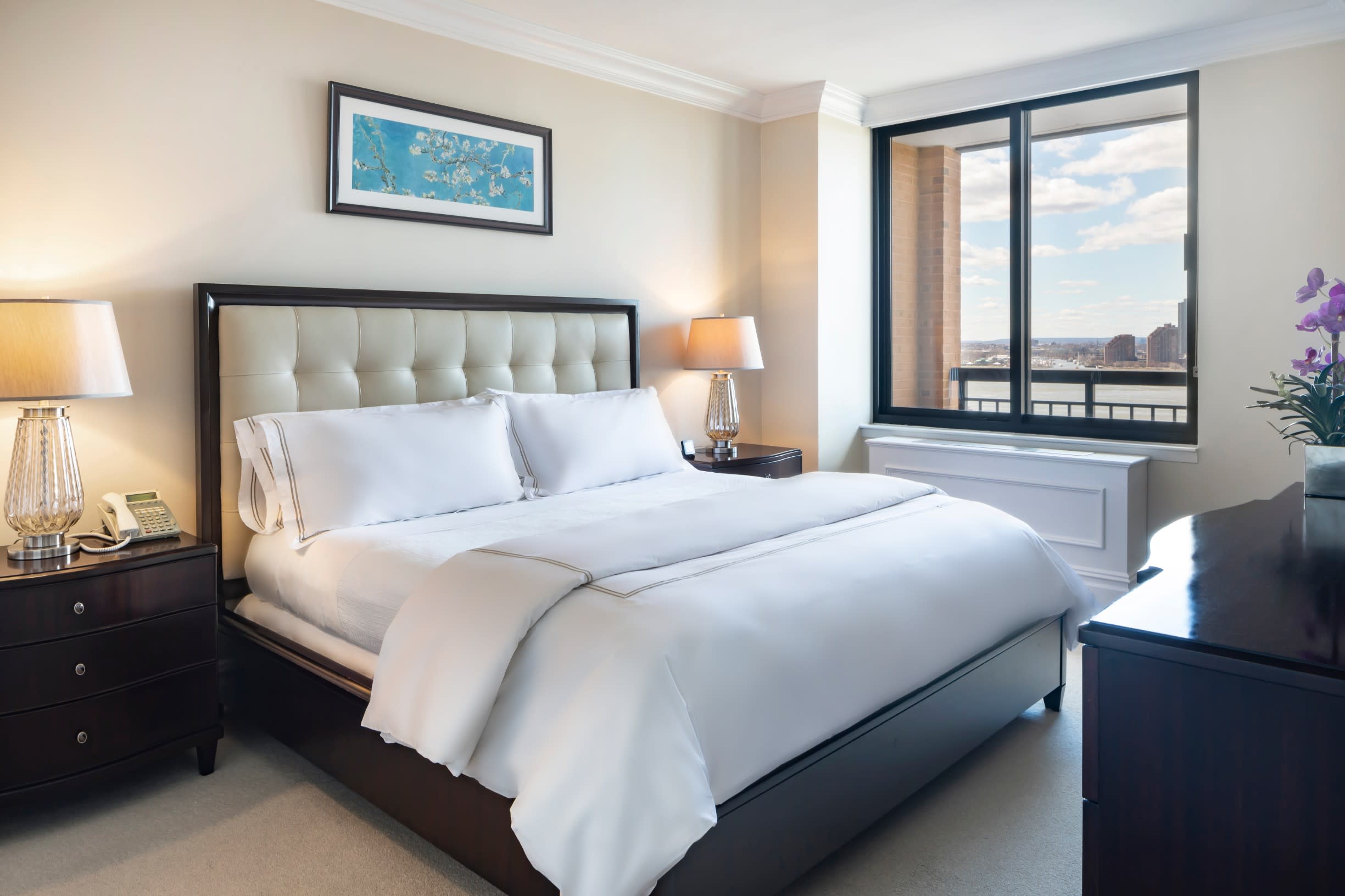 Master bedroom with upholstered headboard, white bedding, dark wood nightstands with lamps, and large windows overlooking the city skyline.