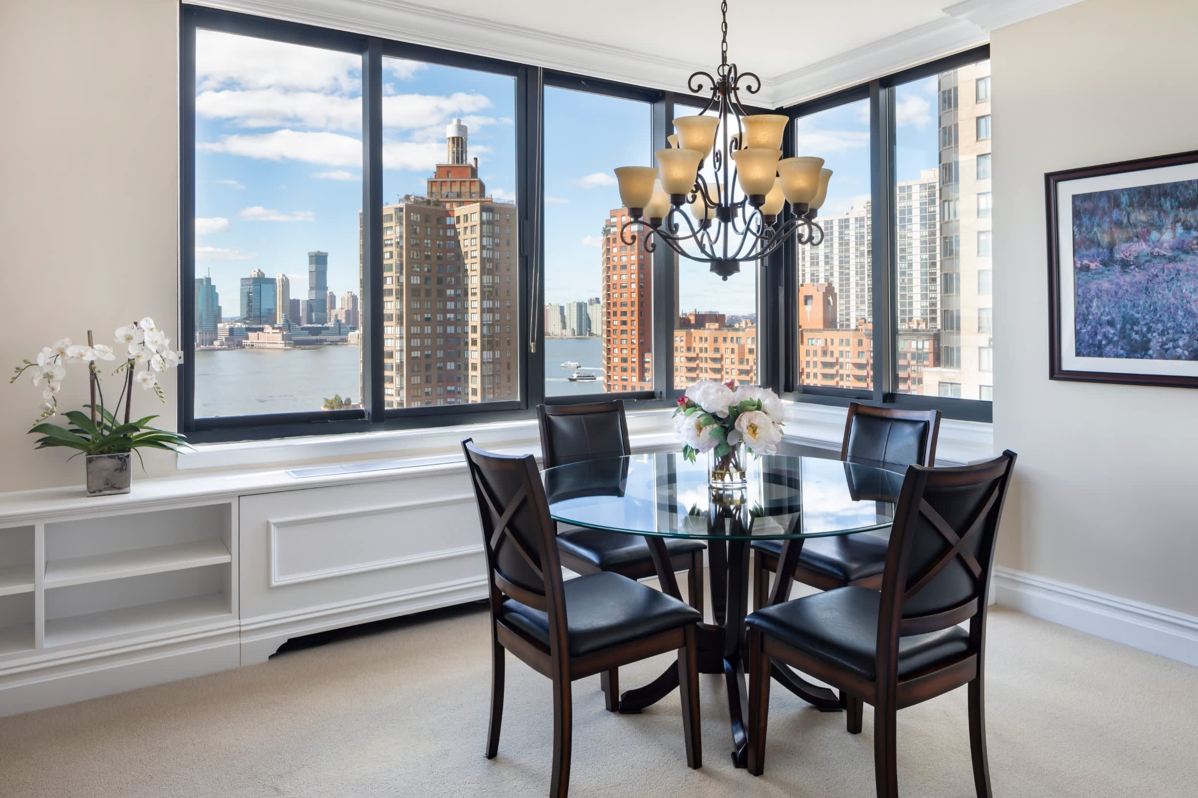 Dining room with round glass table, four black chairs, and expansive windows overlooking Manhattan skyline and East River.
