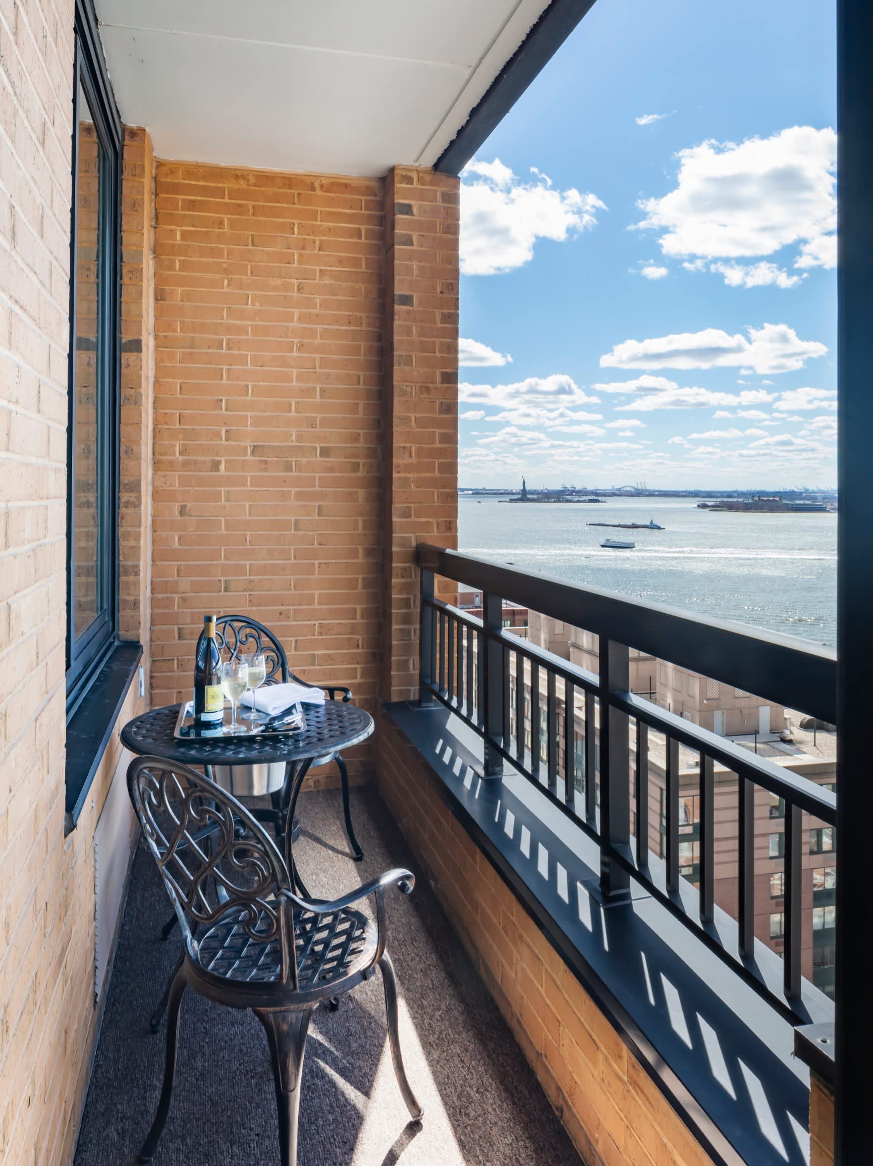 Private balcony with small round table, two ornate chairs, and expansive view of New York Harbor with Statue of Liberty visible in the distance.