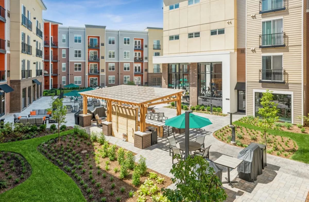 Apartment complex courtyard with wooden pergola, turquoise umbrella, patio seating, and surrounding multi-story residential buildings.