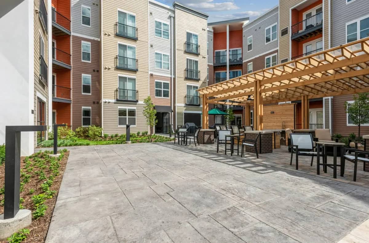 Outdoor courtyard with gray paved flooring, wooden pergola structure, seating areas, and multi-story apartment buildings with red and tan siding.