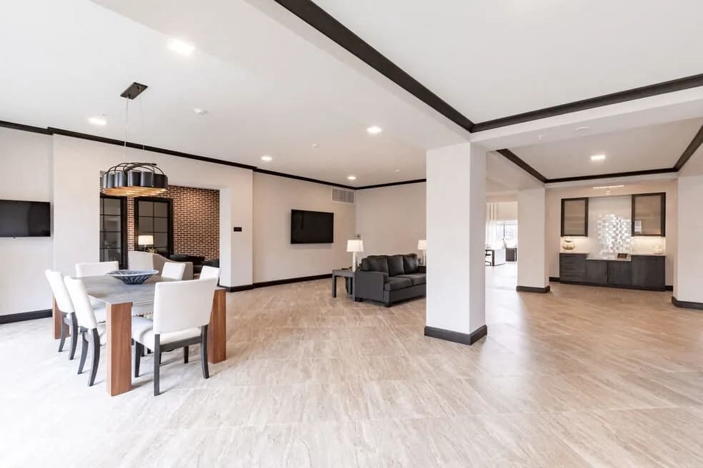 Open-concept apartment interior with wood dining table, white chairs, dark sofa, wood-look flooring, and black accent beams on white ceiling.