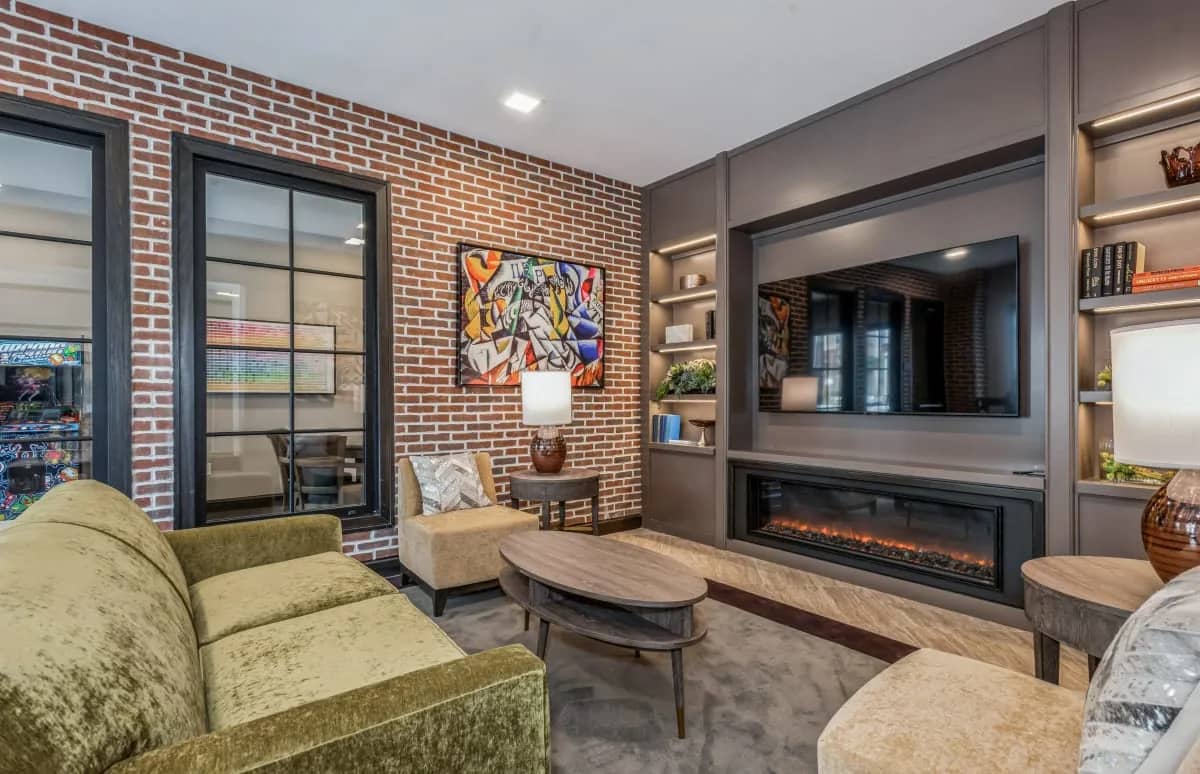 Living room with green velvet sofa, exposed red brick wall, wall-mounted TV above electric fireplace, and gray built-in shelving with ambient lighting.