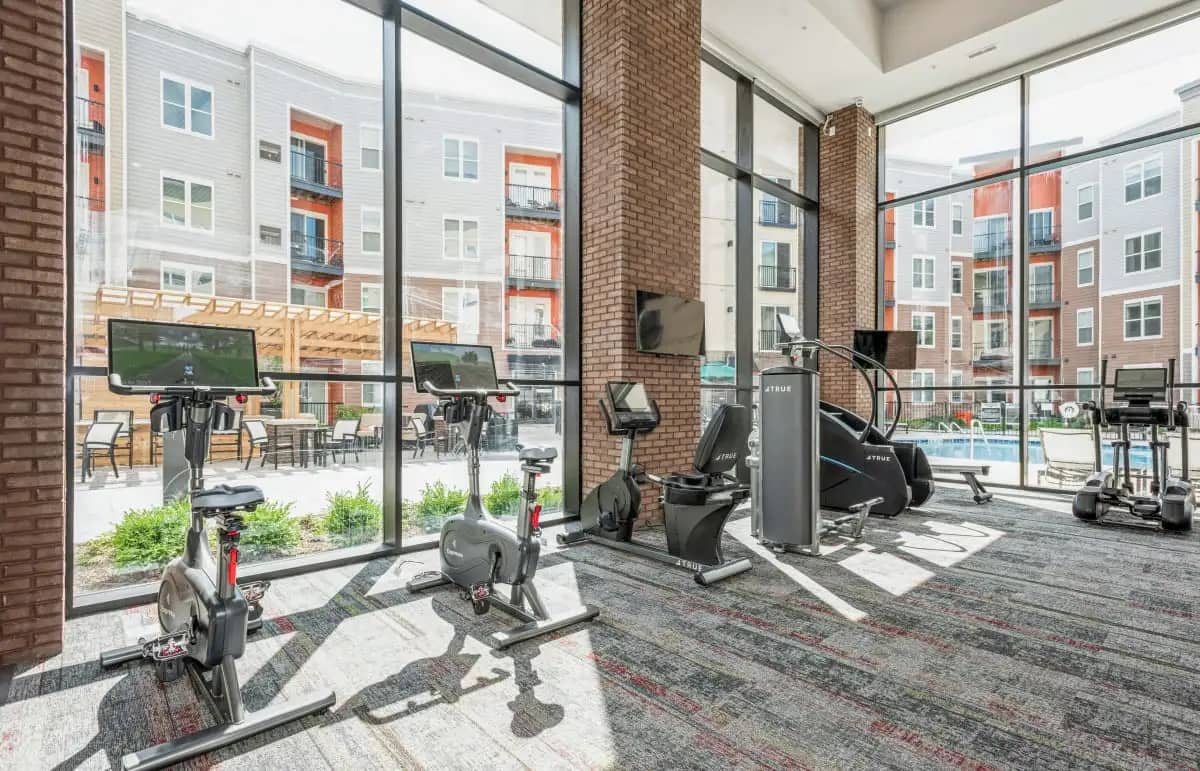 Fitness center with stationary bikes, elliptical machines, and strength equipment; large windows overlook a brick courtyard with pool and residential buildings.