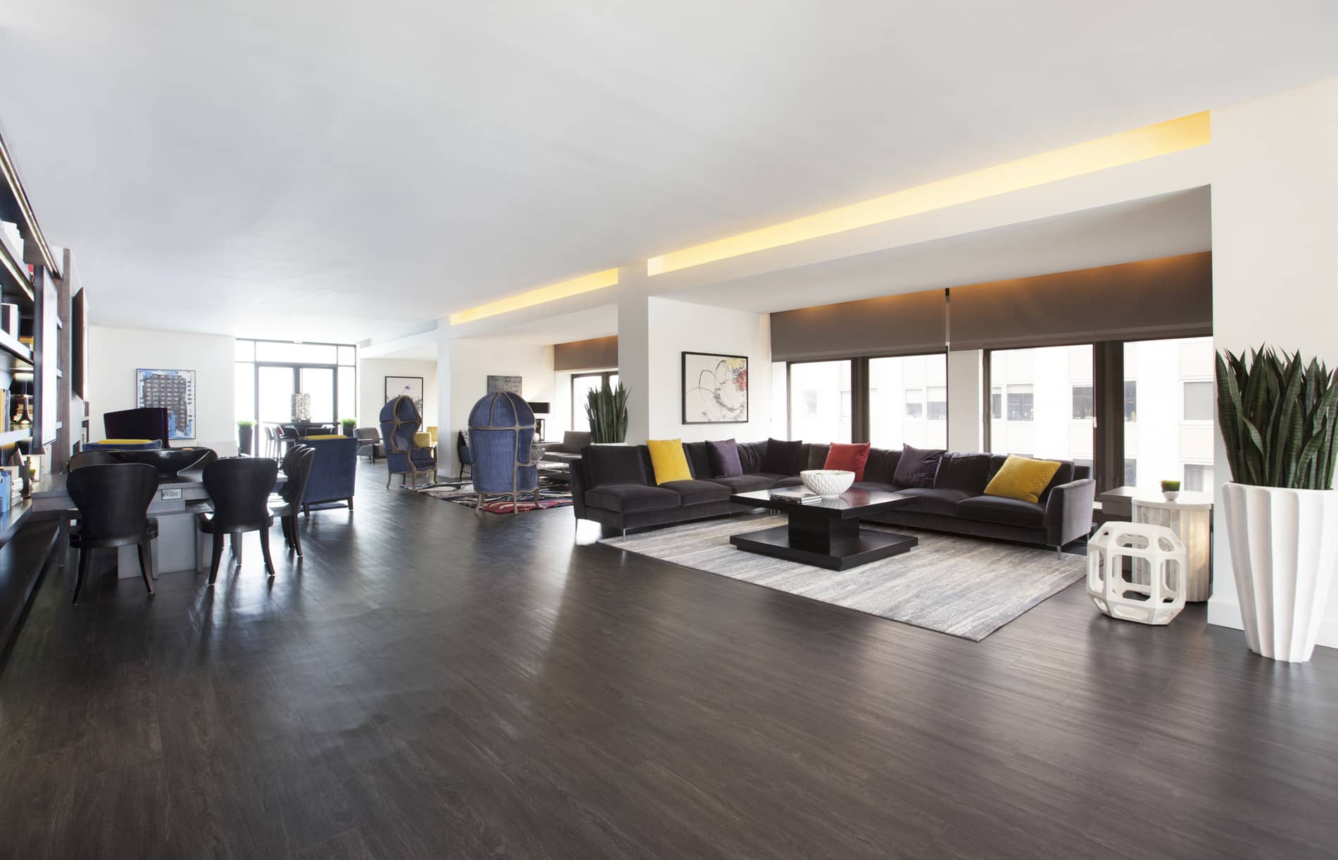 Modern living room with dark wood flooring, black L-shaped sectional sofa with colorful pillows, large windows, and contemporary furnishings.