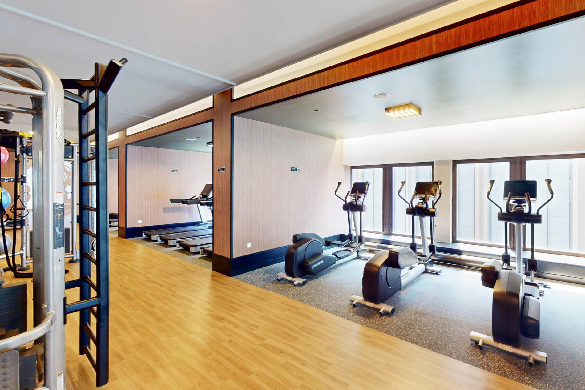 Fitness center with treadmill, stationary bikes, elliptical machines, dumbbells, and mirrored walls on wooden flooring.