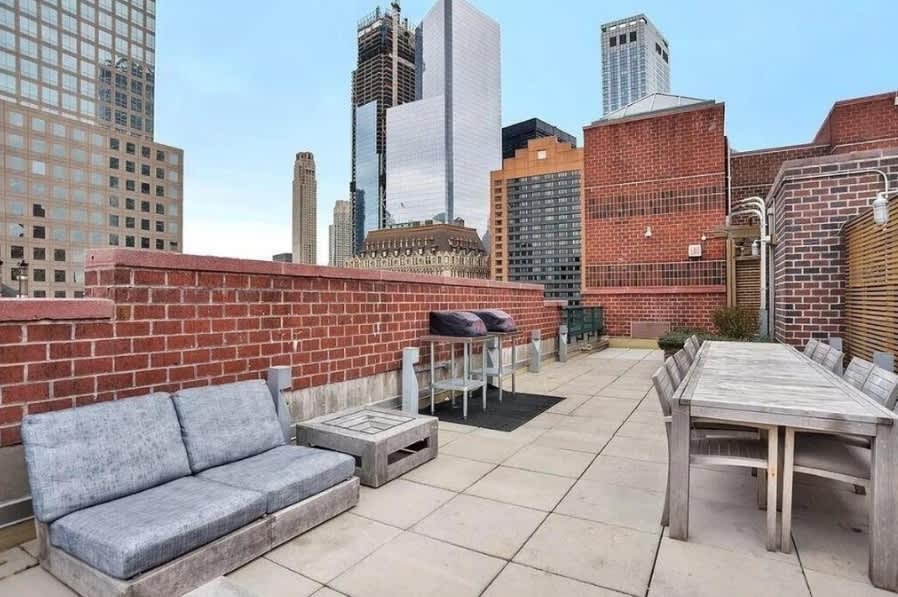 Rooftop terrace with gray seating, wooden dining table, brick walls, and downtown Manhattan skyline views.