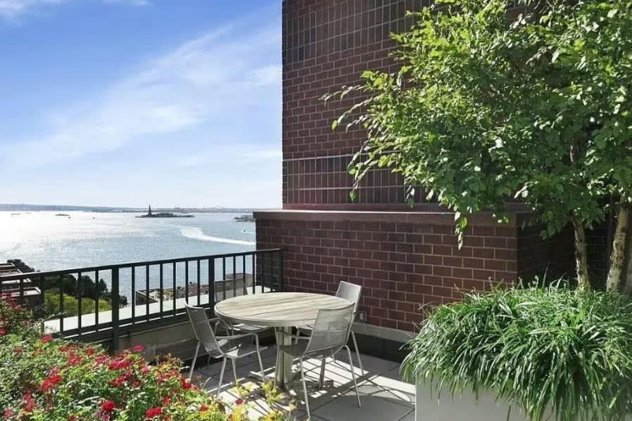 Brick building terrace with round dining table, two chairs, flowering red plants, climbing ivy, and water views beyond black railing.