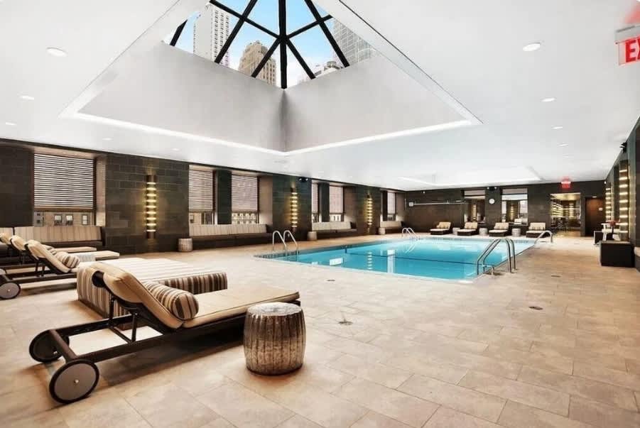 Indoor swimming pool with skylight ceiling, lounge chairs with striped cushions, dark wood walls, and geometric skylight framing city views.